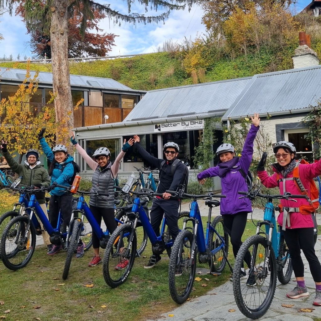 Group of cyclists cheer beside their bikes on a grassy area with autumn trees.