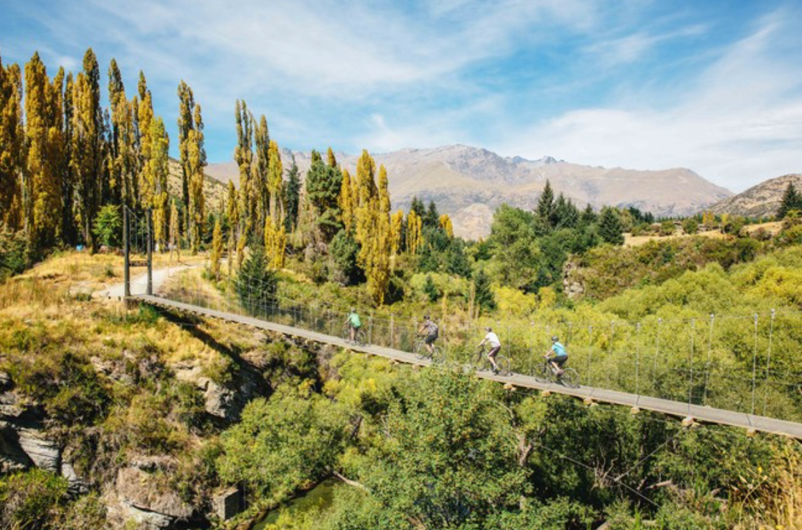 Four cyclists on a bridge with trees and mountains in the background.