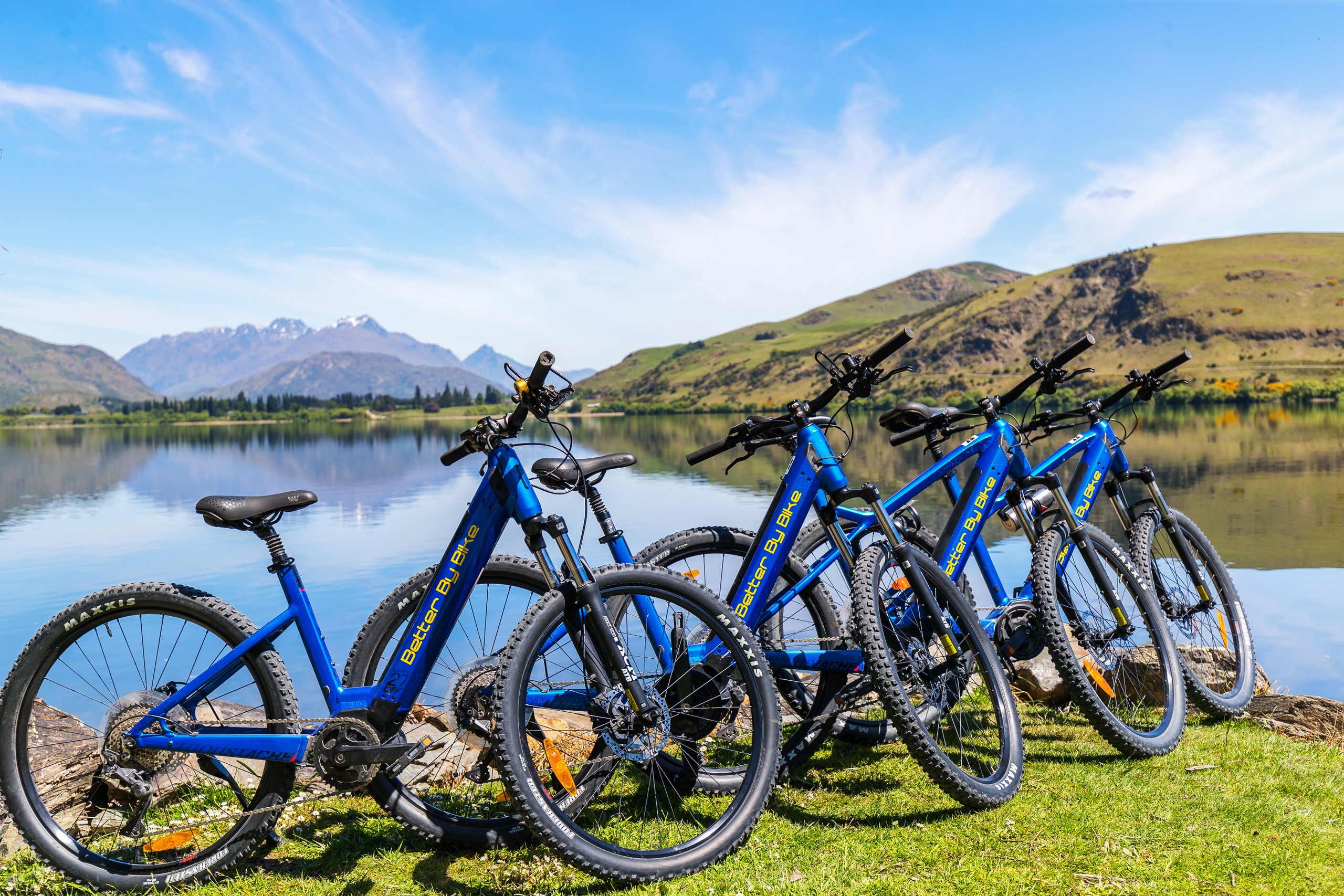 Four blue bicycles parked on grass by a lake with mountains in the background.