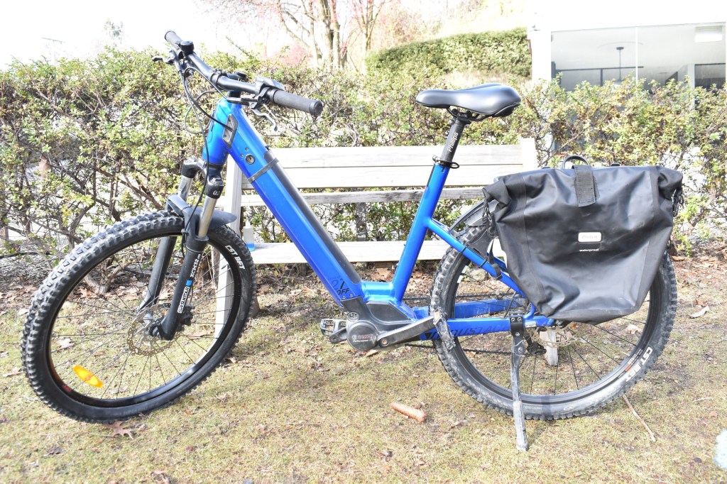 Blue bicycle with a black bag parked near a bench on grass.