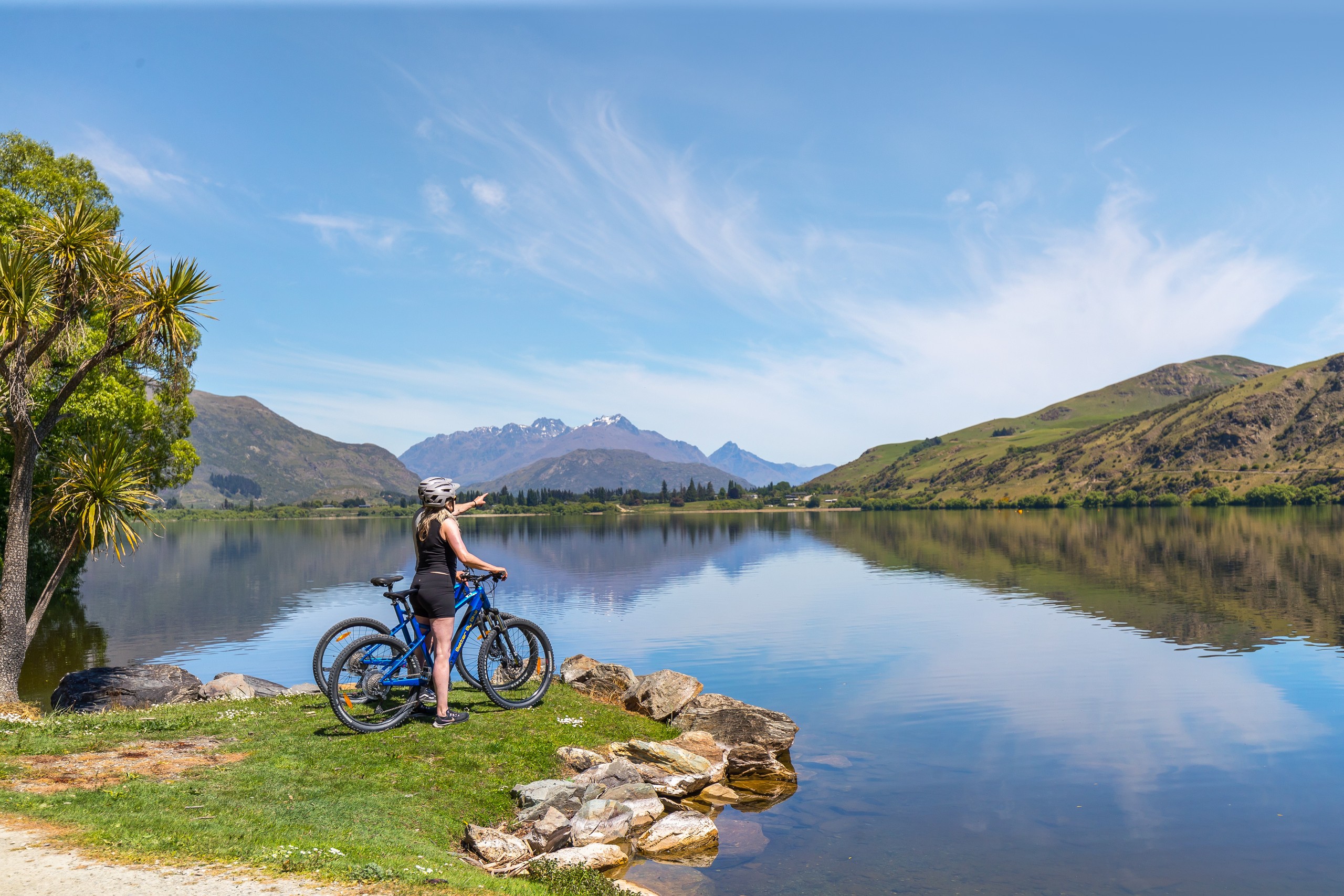 Person with bicycle overlooks a calm lake, mountains in the background, clear sky.