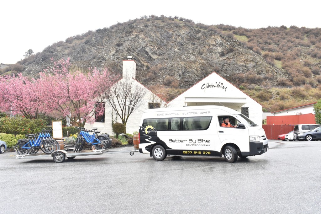 Van with bike trailer parked near building, blooming trees, and rocky hillside.