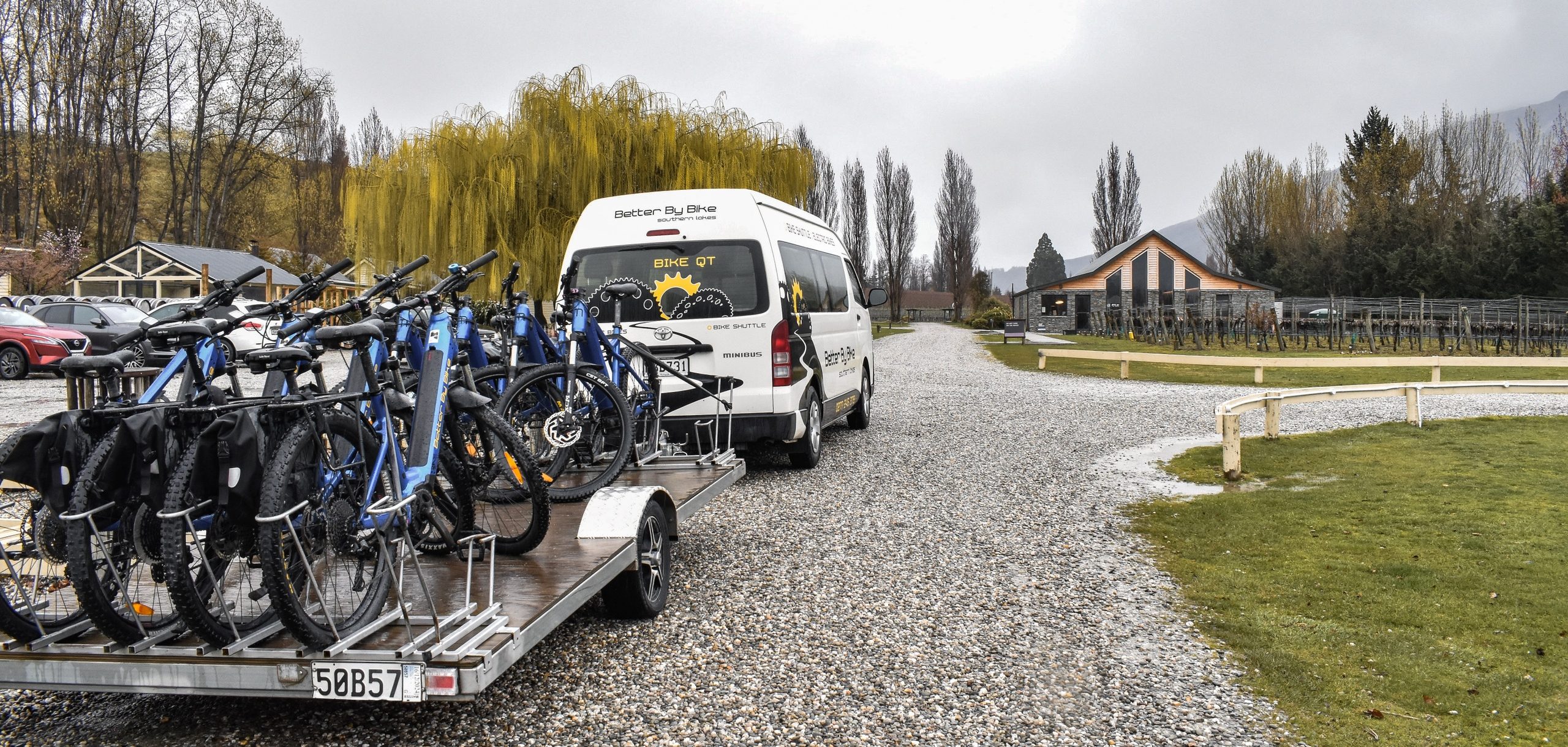 Van towing a trailer with bicycles parked on a gravel path beside a grassy area and trees.