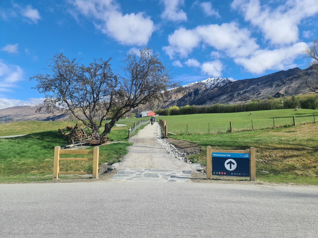 Pathway leading to mountain with tree and grassy fields under a blue sky with clouds.