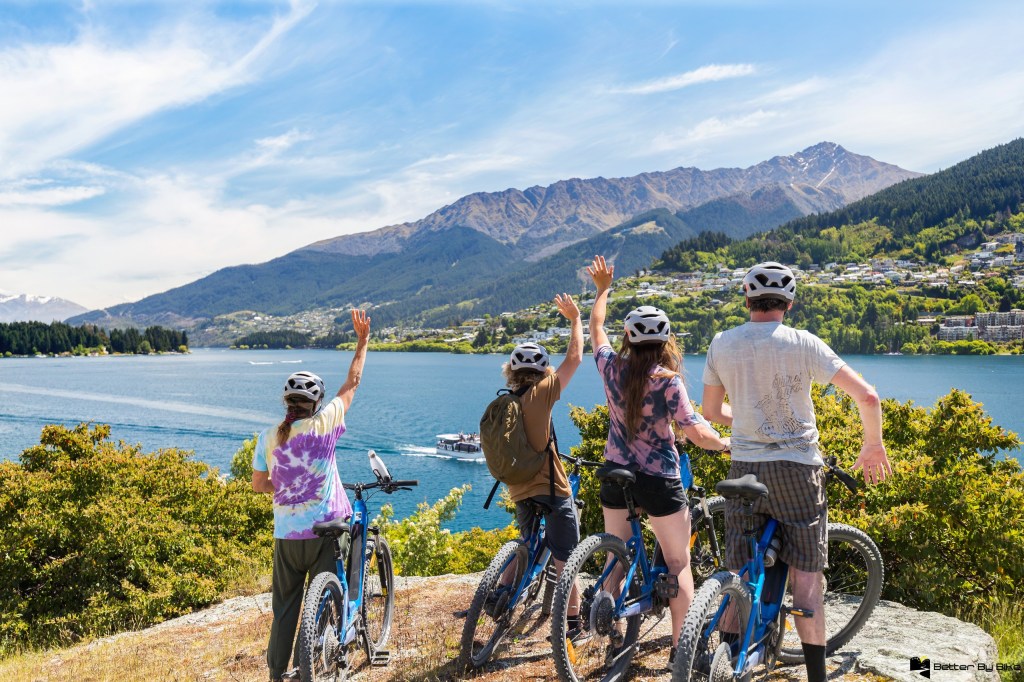 Four people with bicycles facing a lake and mountains, waving.