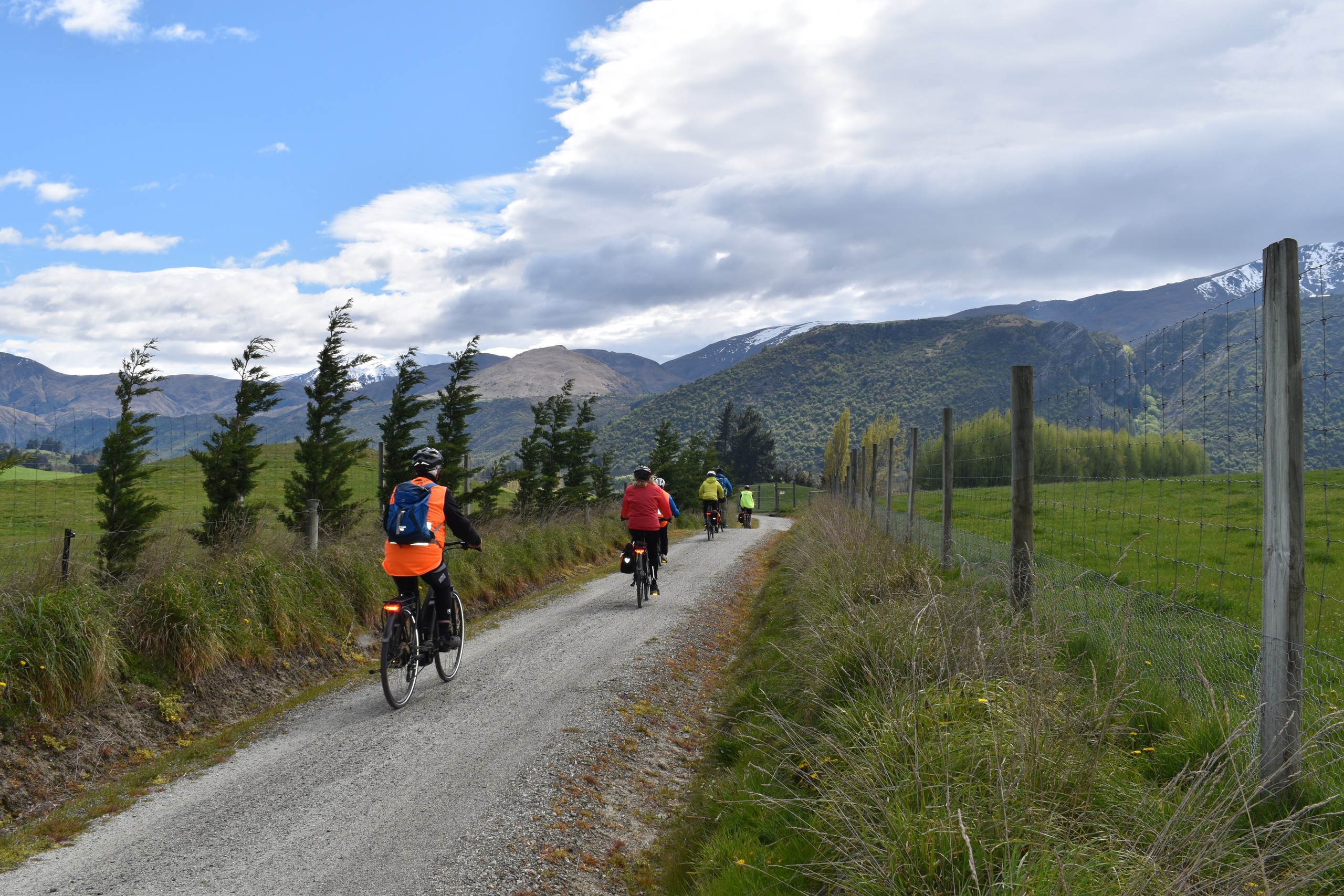 Cyclists on a rural path with mountains and cloudy sky in the background.