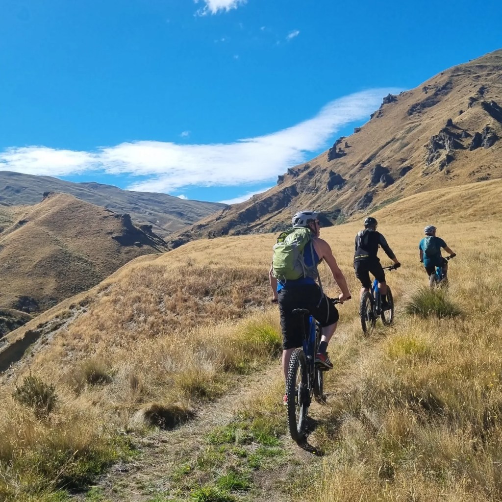 Three cyclists ride on a mountain trail under a clear blue sky.